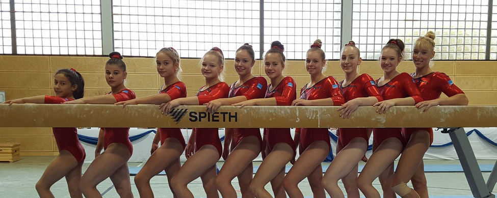 Elf Turnerinnen in roten Trikots am Balancebalken SPIETH