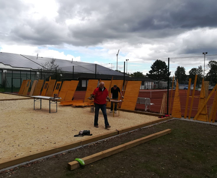 Zwei Männer montieren Holzpaneele an einem eingezäunten Sportplatz