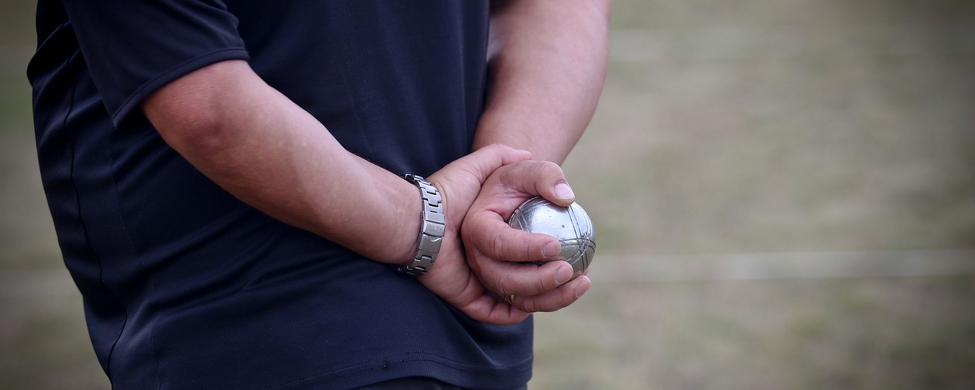 Person hält Boule-Kugel hinter dem Rücken auf Spielfeld