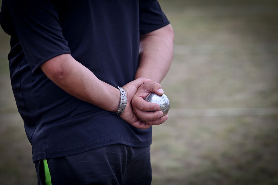 Person hält Boule-Kugel hinter dem Rücken auf Spielfeld