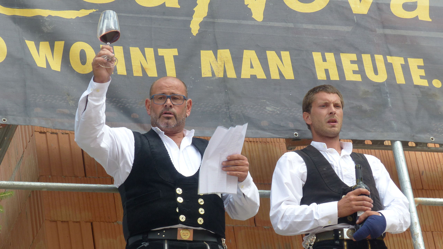 Zwei Männer auf Gerüst, einer hebt Glas, anderer hält Flasche, bandagierte Hand